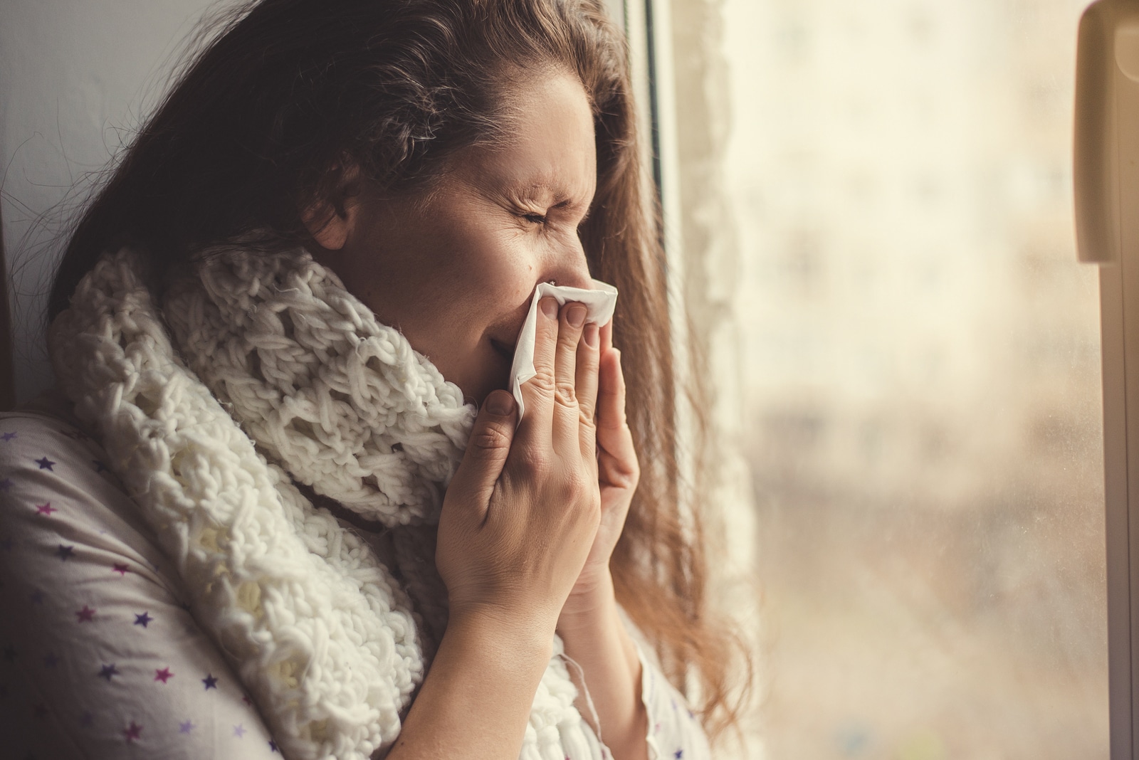 Young girl with a scarf around his neck standing at the window of the patient. Sick girl at the window with a handkerchief.