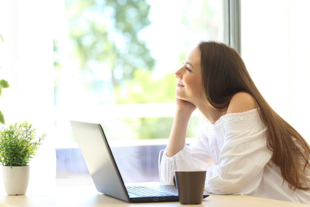 Pensive girl with a laptop daydreaming and looking through a window in a house with a green background outdoors in a beautiful day