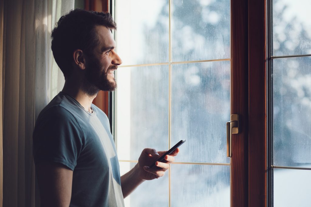 Young bearded man standing by the window using a smartphone