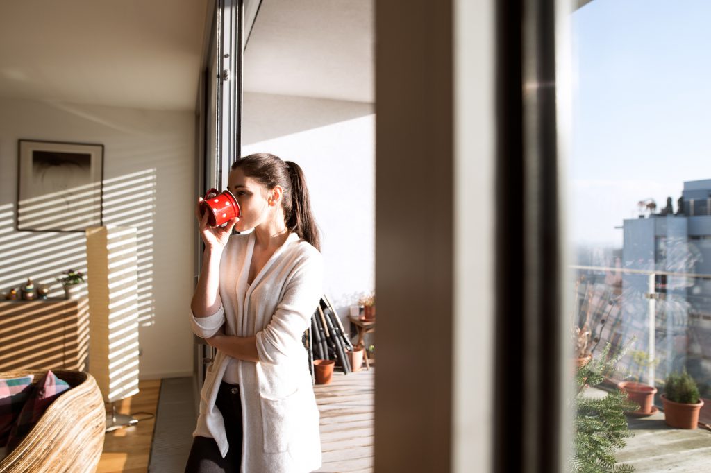 Beautiful young woman relaxing on balcony with city view holding cup of coffee or tea, drinking it