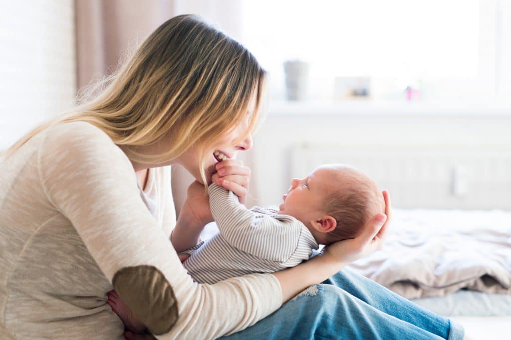 Beautiful young mother with her newborn baby son, sitting on bed in her bedroom, caressing him