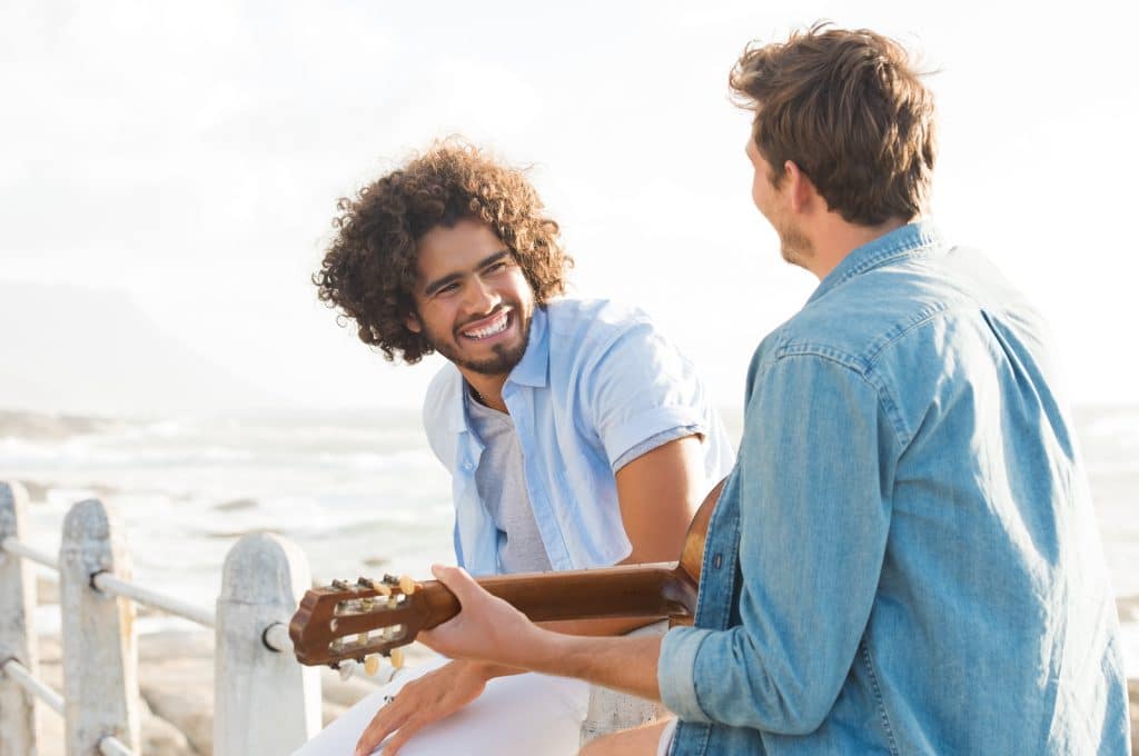 Rear view of young man playing guitar sitting on fence at beach with friend. Smiling guy listening his friend playing acustic guitar at sunset.