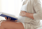 Mulher grávida sentada lendo um livro com a mão sobre a barriga.