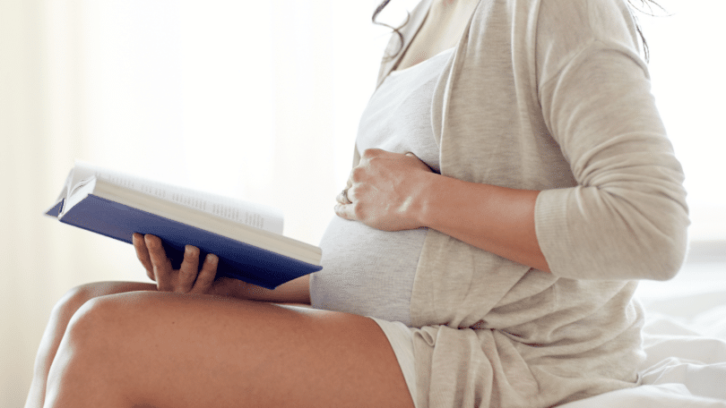 Mulher grávida sentada lendo um livro com a mão sobre a barriga.