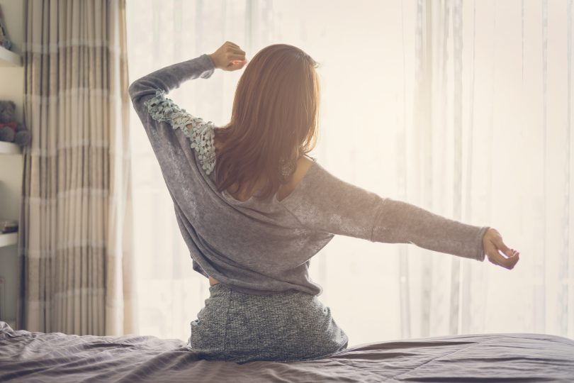 Mulher sentada em cama esticando os braços como se estivesse se espreguiçando.