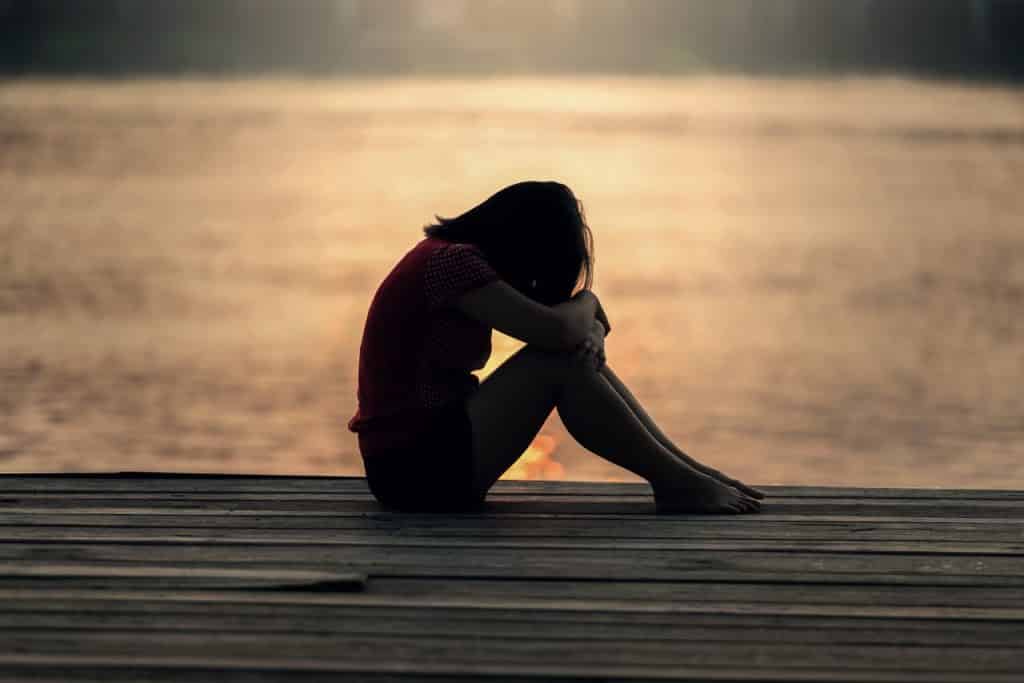 Menina sentada no chão com a cabeça deitada nos braços cruzados que estão apoiados no joelho com o pôr do sol ao fundo.