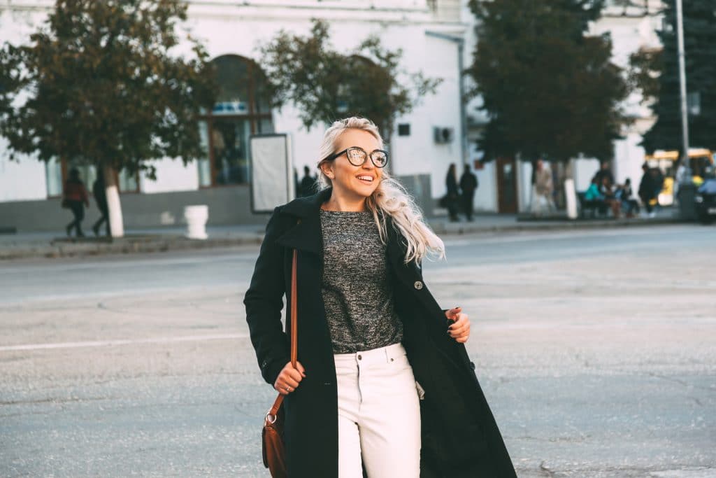 Mulher em pé, sorrindo. Ela está com uma calça branca, uma blusa cinza e um sobretudo preto.