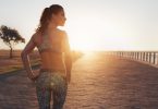 Mulher com roupa de ginástica em calçadão à beira-mar, com o sol no horizonte.