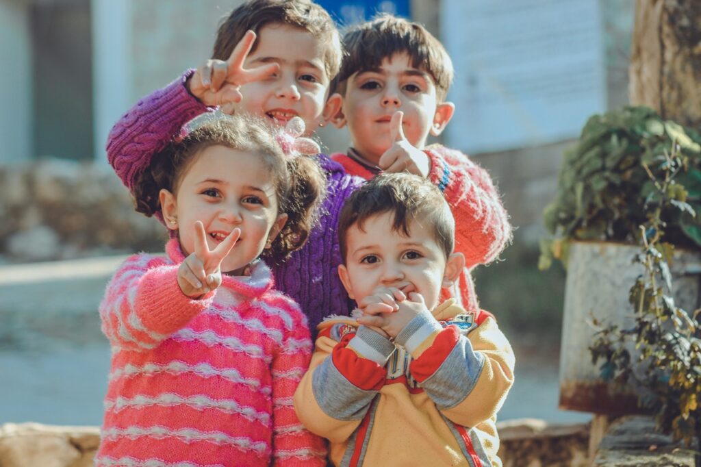 Três meninos e uma menina, com blusas de inverno, sorrindo para a câmera e fazendo gestos com as mãos.