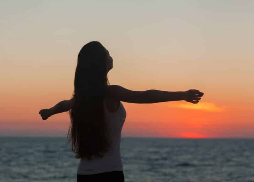 Mulher de braços abertos sob o pôr do sol na praia.