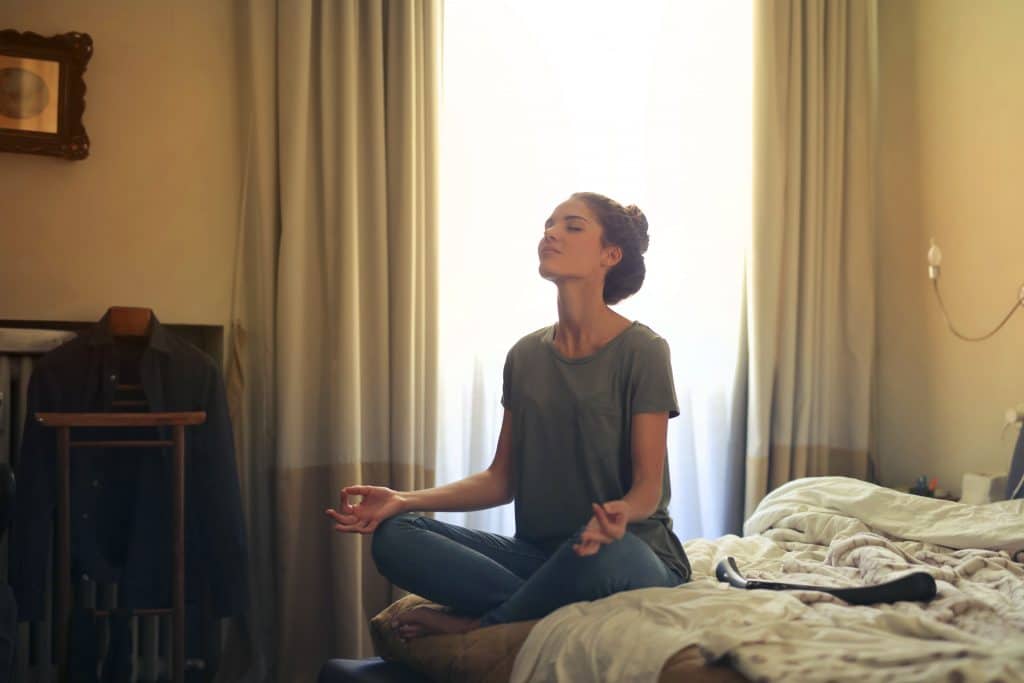 Mulher sentada na cama do quarto, de olhos fechados, meditando.