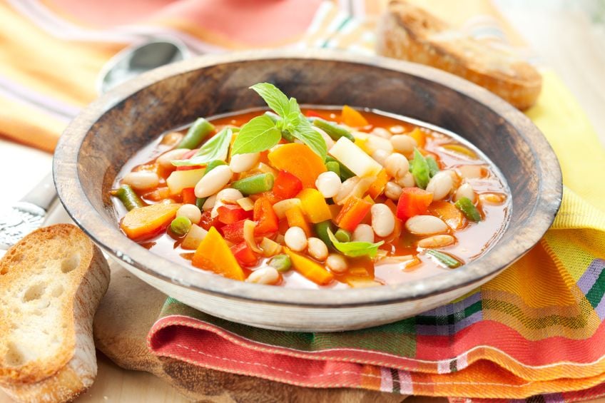 Sopa de lentilha e legumes em um prato fundo de madeira, sobre uma mesa com toalha e torradas.