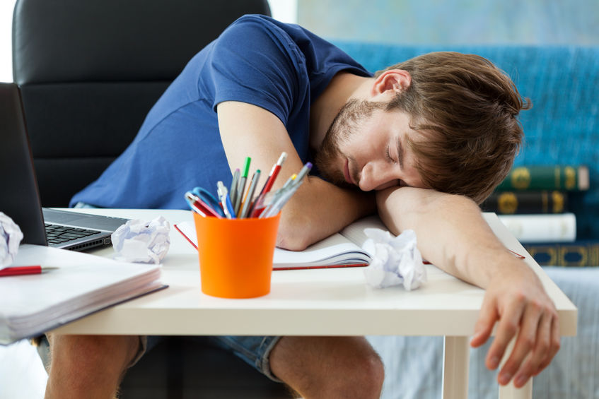 Homem com a cabeça apoiada em uma mesa, sobre seus braços e um caderno, dormindo.