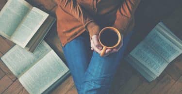 Mulher sentada no chão, com as pernas esticadas, segurando uma caneca com café. Ao seu redor, três livros estão abertos, também no chão.