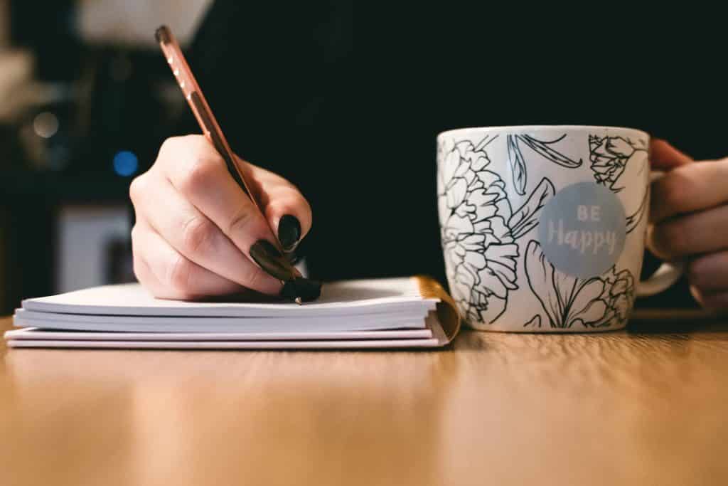 Mulher escrevendo em uma agenda enquanto segura uma caneca de café