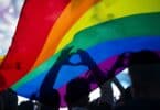 Pessoas debaixo da bandeira LGBTQIAP+ fazendo símbolo de coração com as mãos