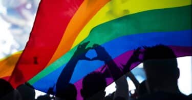 Pessoas debaixo da bandeira LGBTQIAP+ fazendo símbolo de coração com as mãos