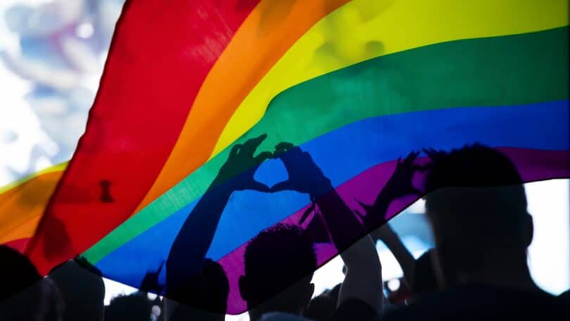 Pessoas debaixo da bandeira LGBTQIAP+ fazendo símbolo de coração com as mãos