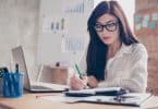 Mulher branca de óculos sentada em frente à uma mesa de escritório escrevendo num caderno.