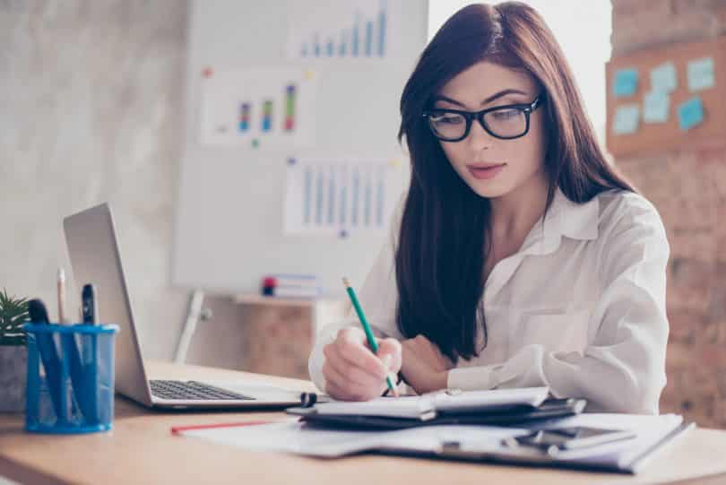 Mulher branca de óculos sentada em frente à uma mesa de escritório escrevendo num caderno.