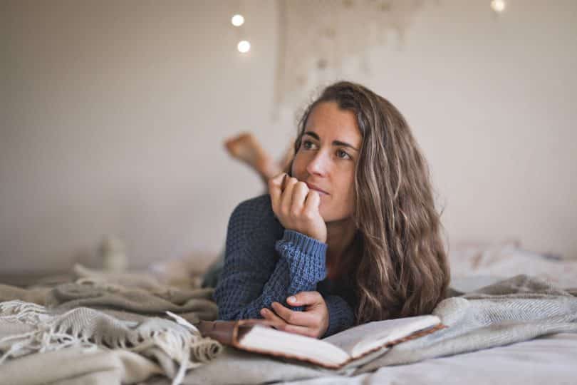 Mulher branca deitada numa cama lendo um livro.