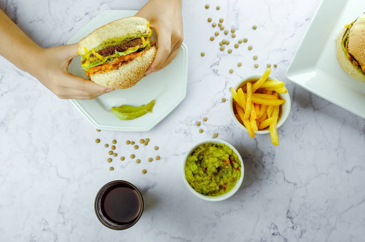 Recorte de uma pessoa segurando um lanche de hambúrguer com aperitivos em volta da mesa.
