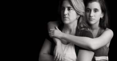 Mãe com expressão séria, de braços cruzados, enquanto sua filha a abraça.