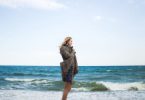Mulher de perfil, em pé, em frente ao mar na praia.