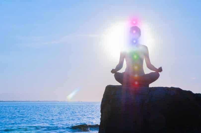 Mulher meditando em cima de uma rocha com a vista para o mar. Os sete chakras estão indicados em seu corpo