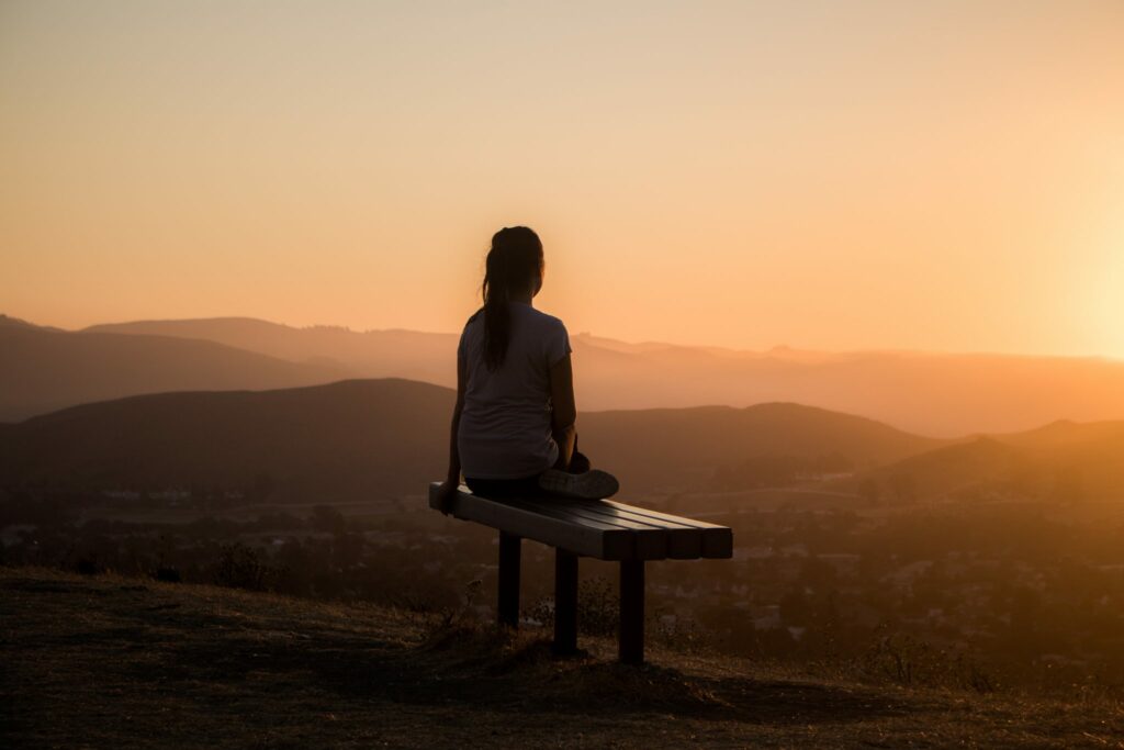 Mulher sentada em um banco observando o pôr-do-sol em um ponto alto de uma cidade.