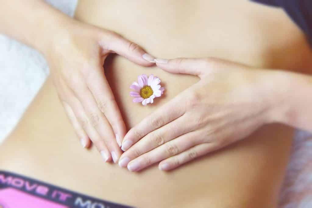 Mulher coloca suas mãos em forma de coração sobre sua barriga, com uma flor ao meio.