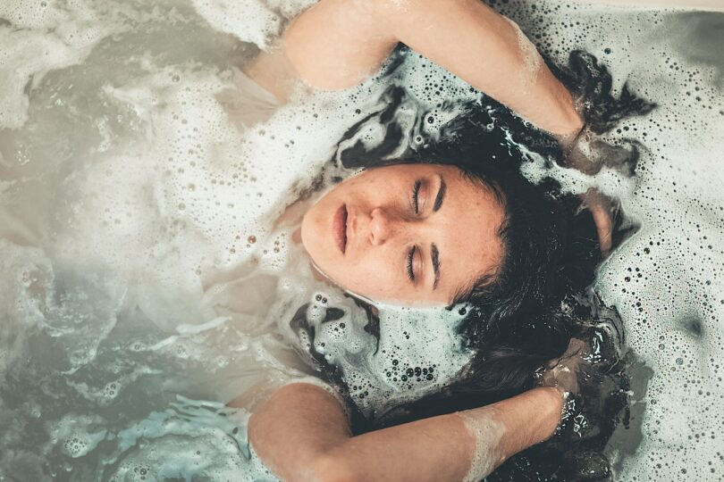 Mulher em uma banheira com espuma, com os braços e o rosto para fora da água.