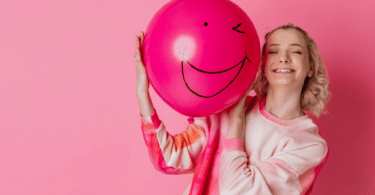 Mulher loira sorrindo enquanto segura um balão rosa com um cara feliz desenhada, em fundo rosa.