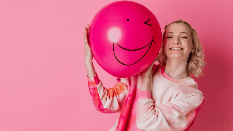 Mulher loira sorrindo enquanto segura um balão rosa com um cara feliz desenhada, em fundo rosa.