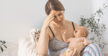 Mãe com a mão sobre a testa, sentindo-se cansada, enquanto bebê chora em querer mamar.