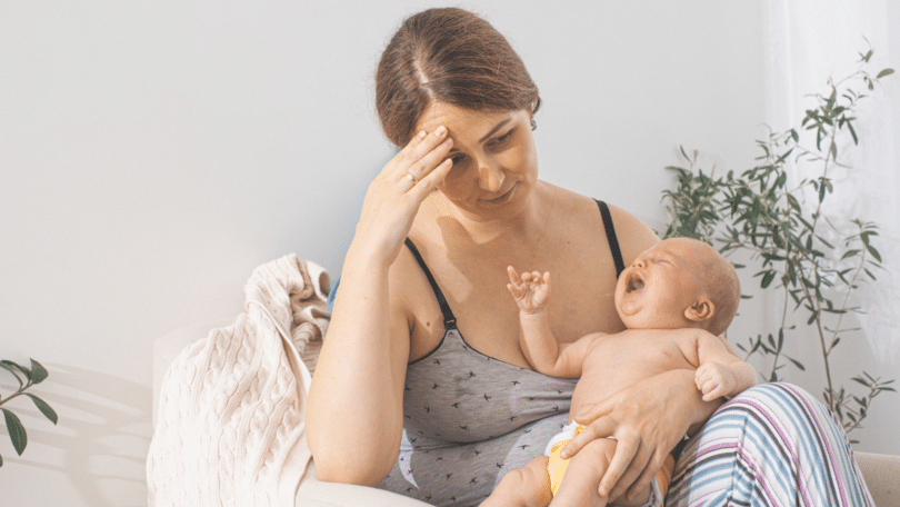 Mãe com a mão sobre a testa, sentindo-se cansada, enquanto bebê chora em querer mamar.