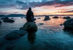 Homem sentado em uma rocha em um mar, olhando para o céu
