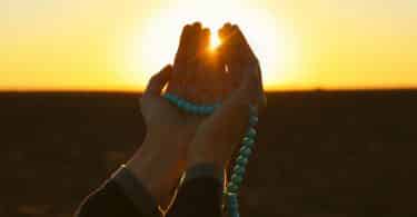 Recorte de uma mão orando com o pôr do sol ao fundo.