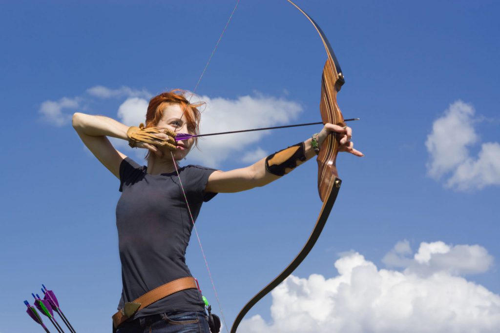 Mulher jovem segurando arco, prestes a soltar uma flecha.