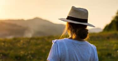 Mulher de chapéu apreciando a vista do pôr do sol sobre uma colina.