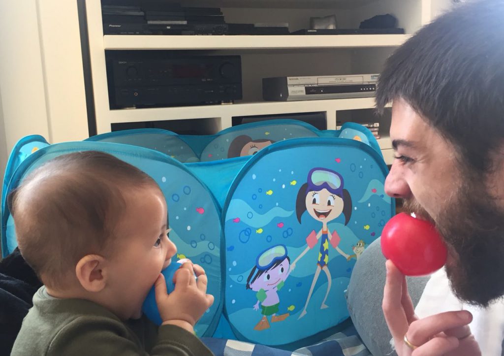 Homem em frente a um bebê colocando uma bolinha de brinquedo na boca