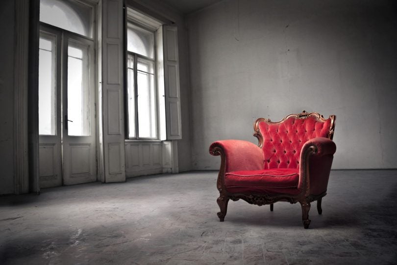 Foto de um amplo cômodo em cor cinza claro, com janelões à esquerda, por onde a luz adentra. Ao lado direito, apenas uma poltrona estilo Luiz XV em madeira entalhada, forrada em veludo vermelho gasto com encosto em capitonê.