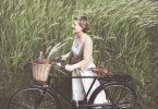 A foto mostra uma mulher sorridente, loira com cabelos curtos lisos e roupas claras leves. Ela está em pé, ao lado de uma bicicleta preta e segura com as duas mãos o guidão. Na cestinha da bicicleta, carrega frutas e uma garrafa. Ao fundo, o vento sopra e inclina a plantação que remete a trigo verde.