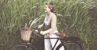 A foto mostra uma mulher sorridente, loira com cabelos curtos lisos e roupas claras leves. Ela está em pé, ao lado de uma bicicleta preta e segura com as duas mãos o guidão. Na cestinha da bicicleta, carrega frutas e uma garrafa. Ao fundo, o vento sopra e inclina a plantação que remete a trigo verde.