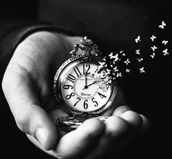 Mão, em preto e branco, segurando relógio que esta desmanchando em fragmentos que se transformam em pequenas borboletas.