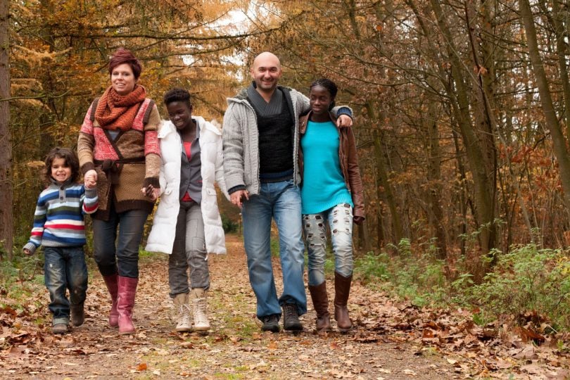 Família formada por um homem e mulher brancos, com 3 crianças negras, caminhando em meio a natureza.