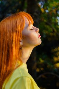 Mulher com cabelo liso e ruivo, de perfil com os olhos fechados, com a cabeça levemente inclinada para cima e com a luz do sol batendo sobre seu rosto.