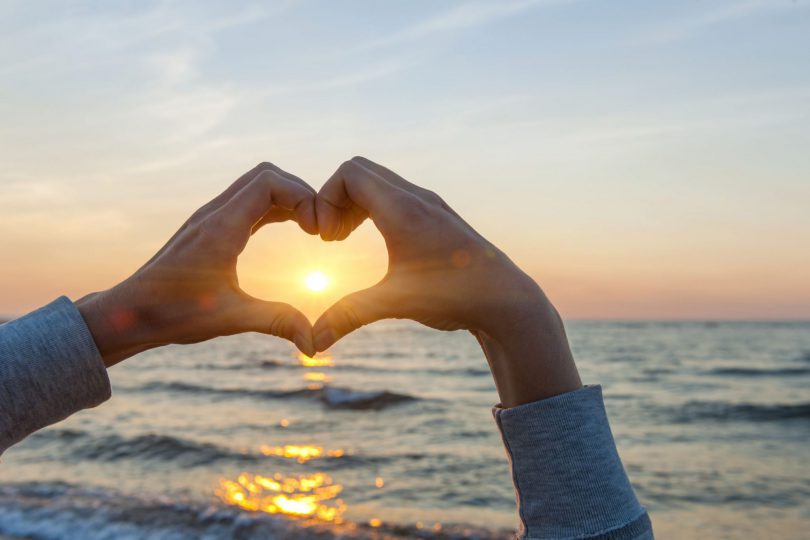 Mãos em formato de coração. Apontadas para o horizonte. Com o pôr do sol e o mar ao fundo.