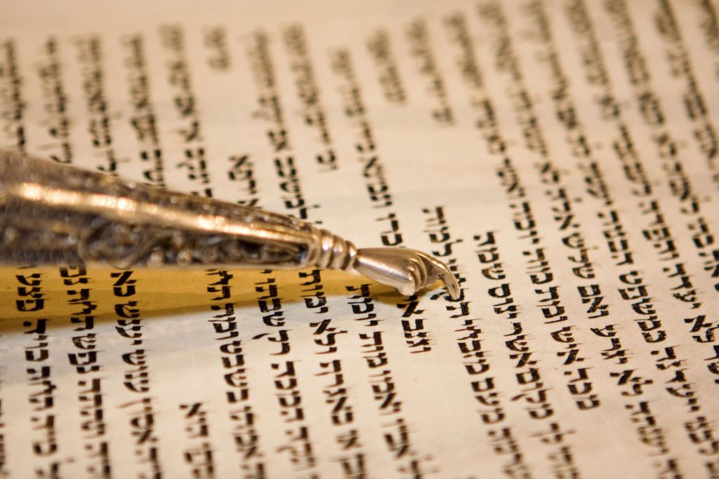 Papel envelhecido com escrituras em hebraico e por cima há um cabo fino e dourado que contém uma pequena mão também dourada que é utilizada para acompanhar a leitura do texto.