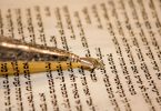 Papel envelhecido com escrituras em hebraico e por cima há um cabo fino e dourado que contém uma pequena mão também dourada que é utilizada para acompanhar a leitura do texto.